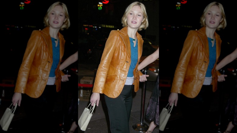 Julia Stiles wearing black trousers, a turquoise top, and a caramel brown leather jacket