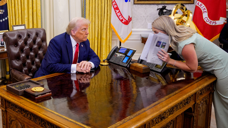 Karoline Leavitt leaning over Donald Trump's desk and hiding her mouth behind a newspaper while speaking to him