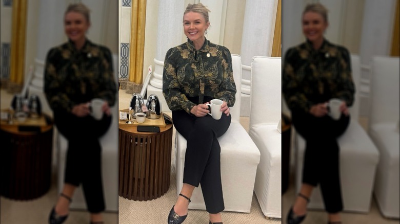 Karolina Leavitt in a pussybow blouse and black slacks, sitting on a white chair