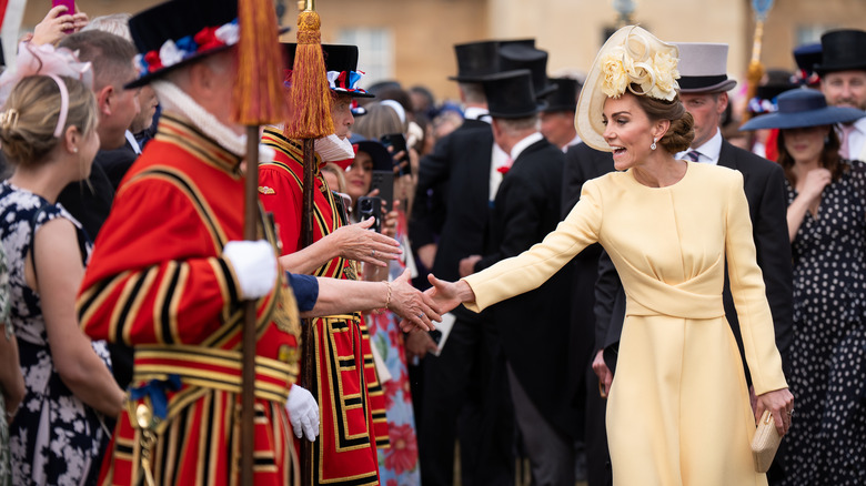 Kate Middleton wearing a yellow dress while reaching to grasp a fan's hand