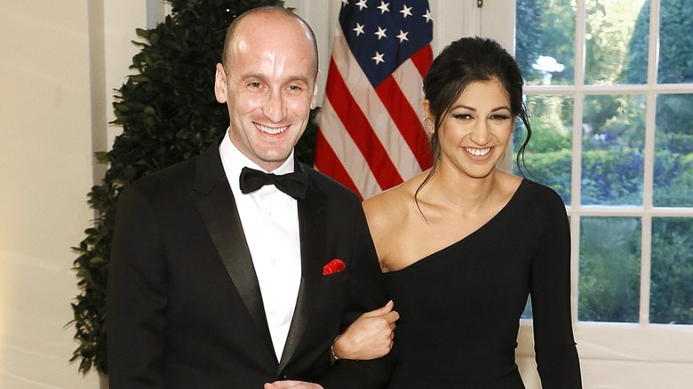Stephen Miller and Katie Miller smiling and walking arm in arm with a U.S. flag in the background