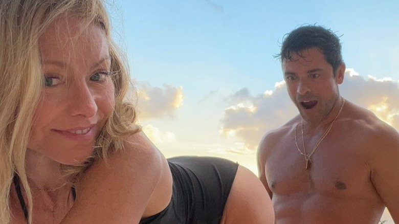 Kelly Ripa in a swimsuit and wearing no makeup bending over in front of Mark Consuelos