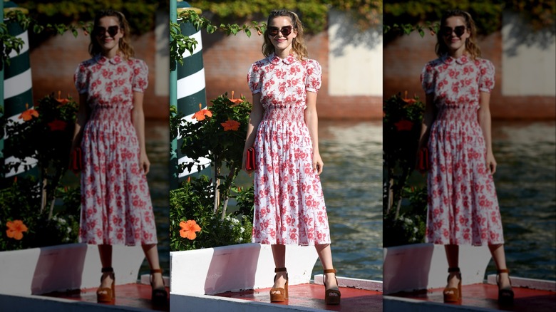 Kiernan Shipka smiling while standing outside in a floral, white-and-red dress with platform heels