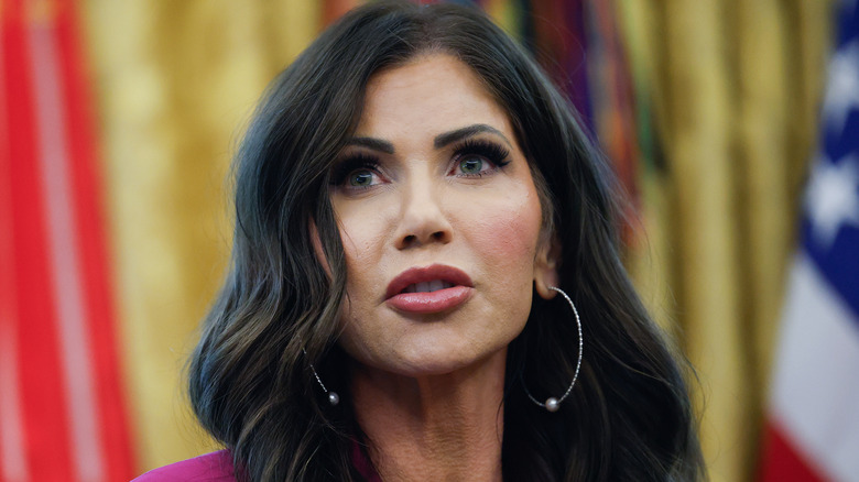 Kristi Noem speaking in the Oval Office in a maroon blazer and silver hoop earrings