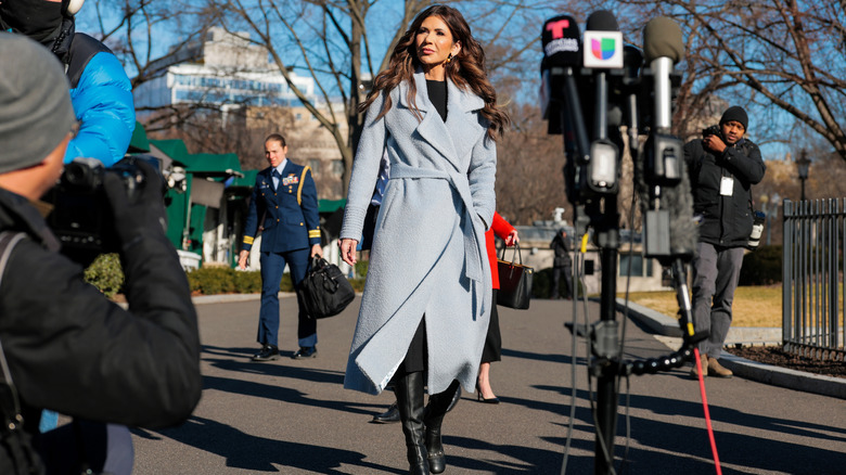Kristi Noem, in long belted coat over black dress and matching boots, walks past reporters outside of the White House in 2026.