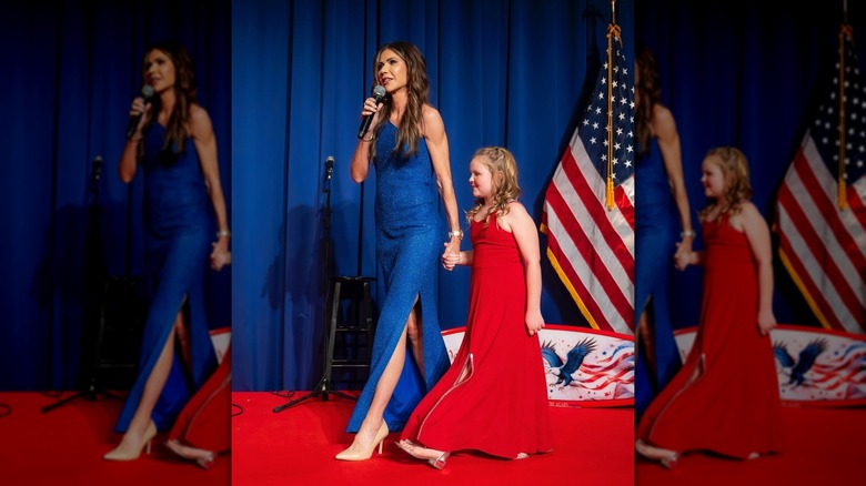 Kristi Noem on stage in a blue gown holding hands with a young girl in a red dress
