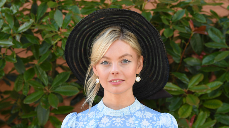 Nell Hudson posing in front of greenery wearing a black hat and blue dress