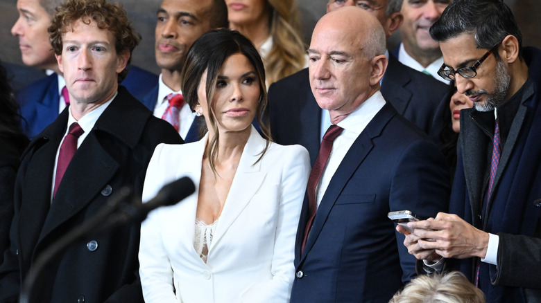 Lauren Sánchez Bezos wearing a white, low-cut suit while flanked by Mark Zuckerberg and Jeff Bezos