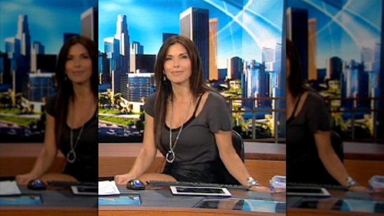 Lauren Sánchez Bezos sitting at a news desk in a gray t-shirt that is nearly slipping off her shoulder, revealing a black strap underneath