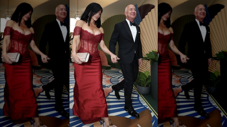 Jeff Bezos walking in hand-in-hand with Lauren Sánchez Bezos who is wearing a red dress with a lace corset bodice