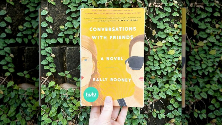 A hand holding up a copy of "Conversations with Friends" in front of an ivy covered wall