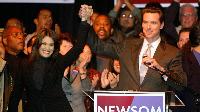 Kimberly Guilfoyle and Gavin Newsom holding hands at a political rally, with supporters in the background