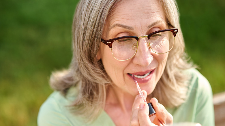 Close up of a mature woman with glasses looking into an unseen compact and applying a lip color