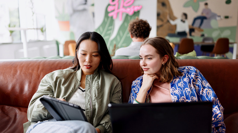 Two women, sitting on a couch, look at a tablet screen