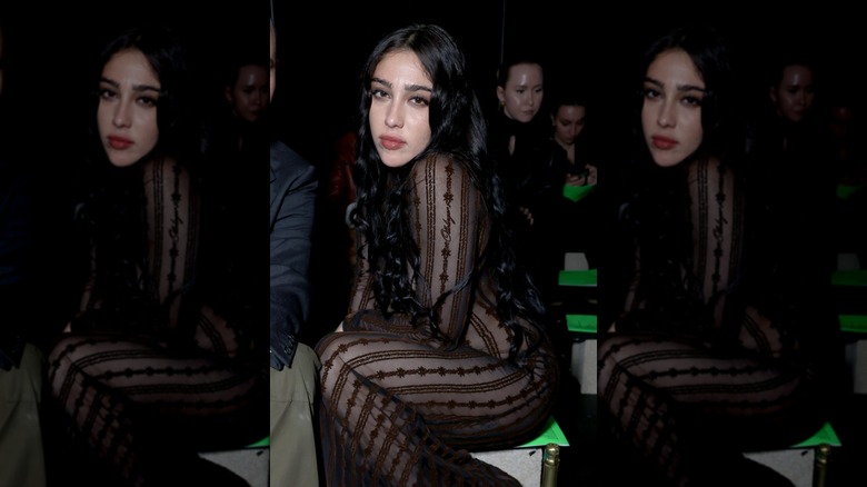 Lourdes Leon sitting down and posing in a sheer, black jumpsuit
