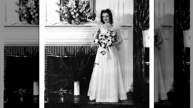 A 16-year-old Marilyn Monroe wearing a wedding dress and holding a bouquet in front of a fireplace