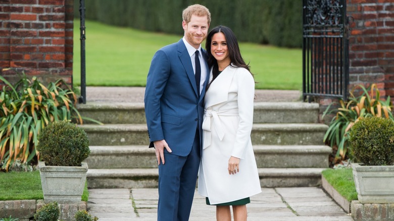 Prince Harry in a suit and Meghan Markle in a fitted, white coat, posing outside