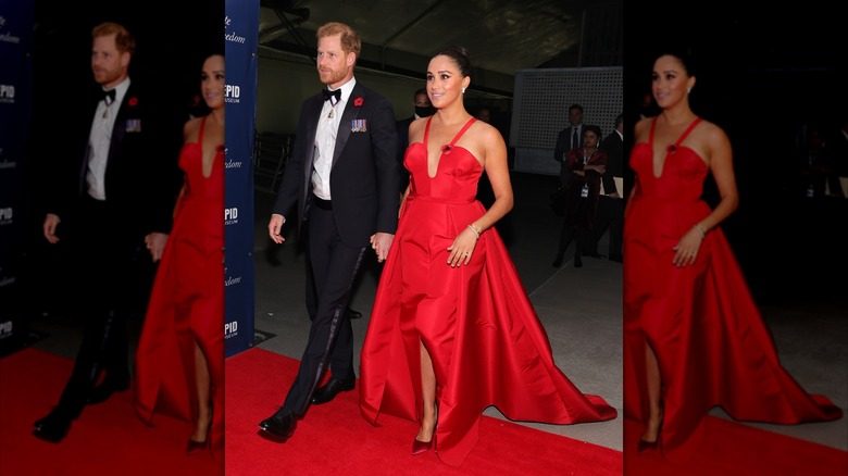 Prince Harry, in a tux, walking hand in hand on the red carpet with Meghan Markle, in a red dress, at the 2021 Freedom Gala