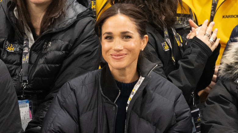 Meghan Markle wearing a puffer jacket and sitting among a crowd