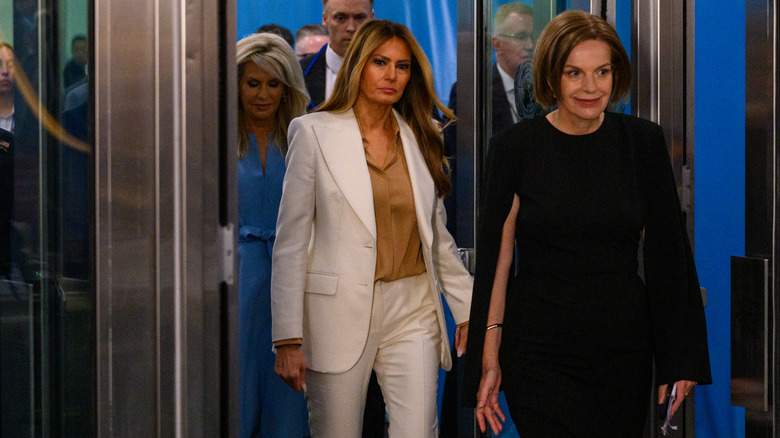 Melania Trump walking through a doorway in a white suit with other women around her