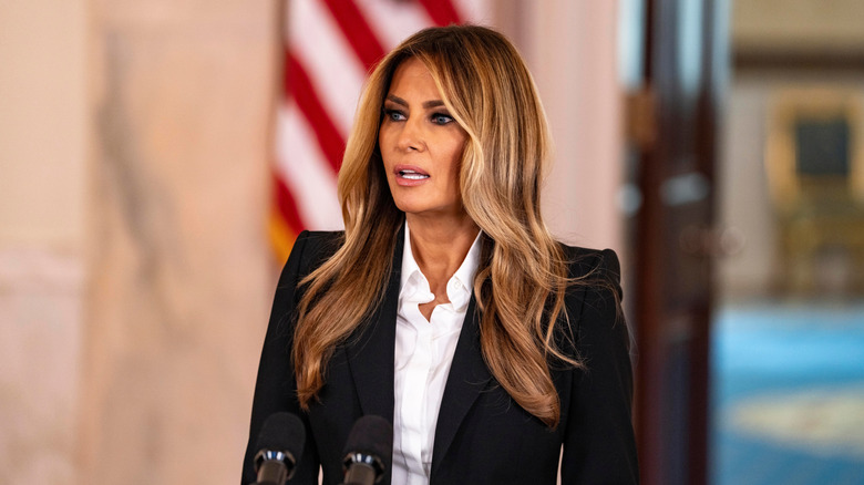 Melania Trump speaking at a podium in a black jacket over a white shirt