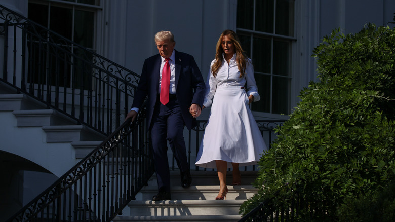Melania Trump walking down exterior steps in a white dress while holding hands with Donald Trump