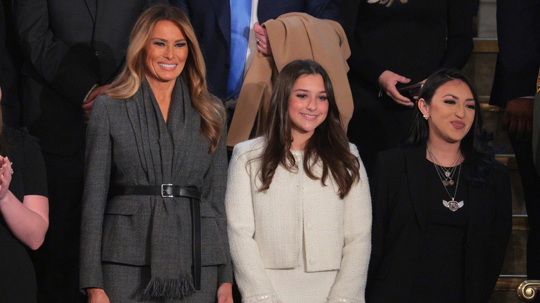 Melania Trump standing and smiling in a grey coat with a scarf