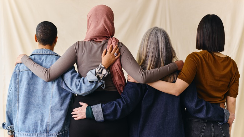 Four women stand facing away from the camera, with their arms around each other