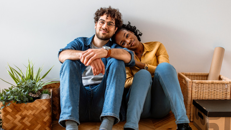Couple happily sitting against a plain wall while surrounded by moving items