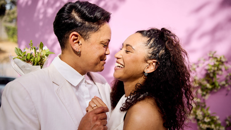 Queer couple smiling as they embrace in white wedding attire in front of a pink building