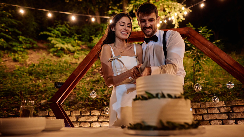 Young newlywed couple cuting a two-tiered cake in front of a geometric, wooden arch decorated with edison bulb lights and mini disco balls