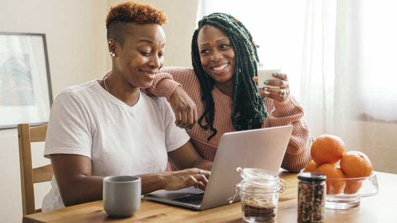 Same-sex couple smiling while sitting at kitchen table and browsing laptop
