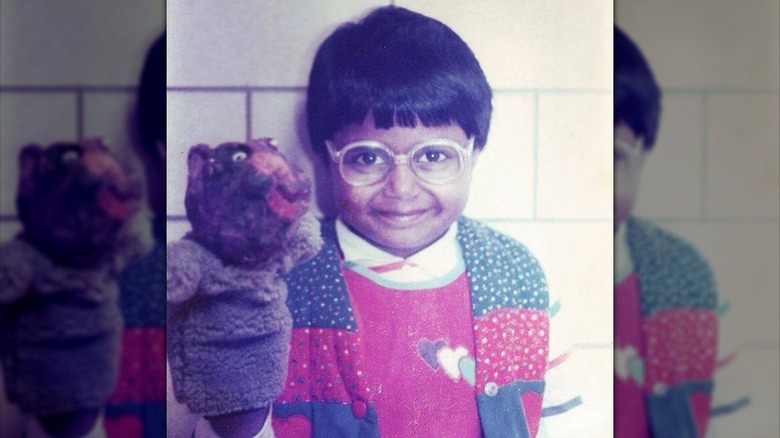A young Mindy Kaling wearing glasses and posing with a plush toy