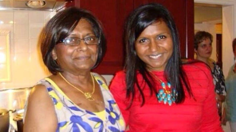 Mindy Kaling, wearing a red dress, smiling next to her mom in a blue-and-yellow dress