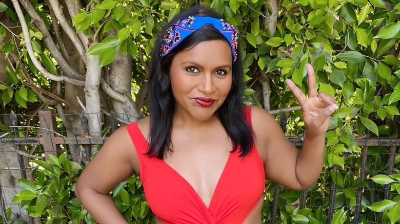 Mindy Kaling posing in an orange swimsuit and flashing a peace sign in front of a leady background
