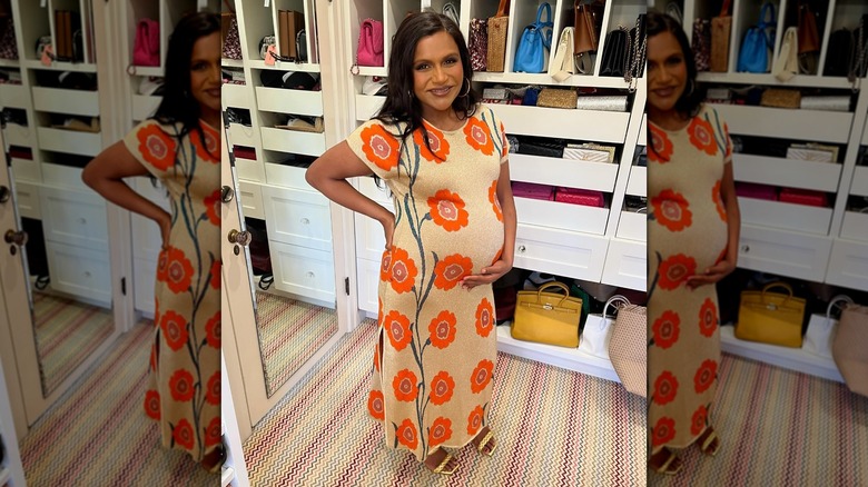 Mindy Kaling cradling her baby bump in an orange, floral dress in 2024