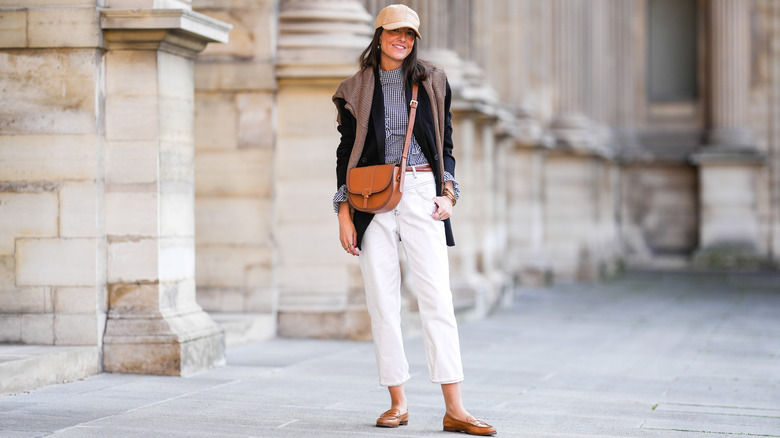 A woman in flats and a cream pair of pants with a jacket and sweater over her shoulders