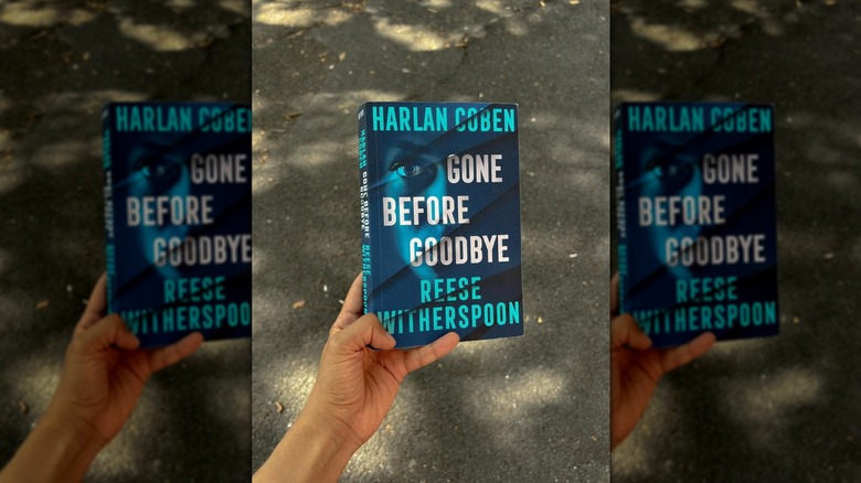 A hand holding a copy of "Gone Before Goodbye" with pavement behind