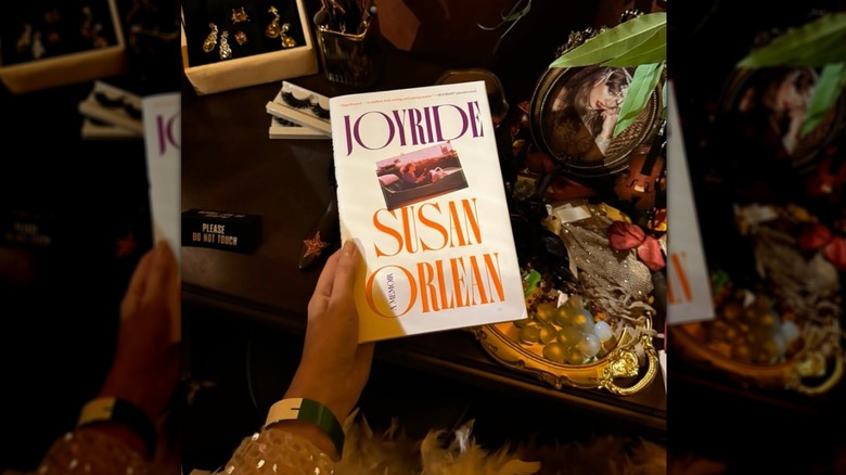 A hand holding the book "Joyride" in front of a table cover in sparkly knickknacks