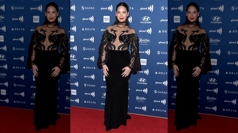Olivia Munn smiling while wearing a gown with peacock silhouettes on the sheer bodice