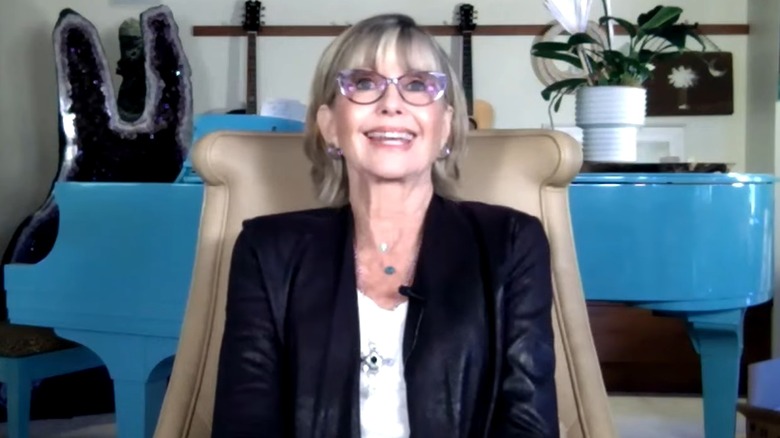 Olivia Newton John sitting in a chair and smiling while wearing a black jacket and glasses