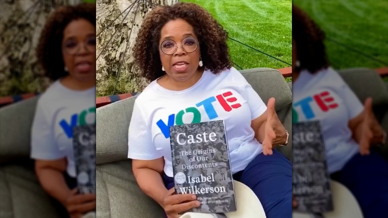 Oprah Winfrey sitting outside and holding a copy of Caste: The Origins of Our Discontents by Isabel Wilkerson,