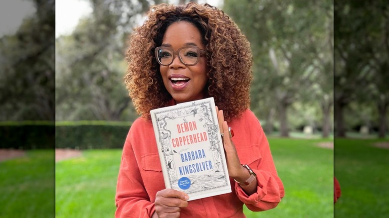 Oprah Winfrey smiling while holding a copy of Demon Copperhead by Barbara Kingsolver