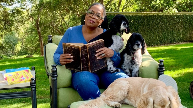 Oprah Winfrey reading a book outside while sitting with her dogs, with books on a table to her right