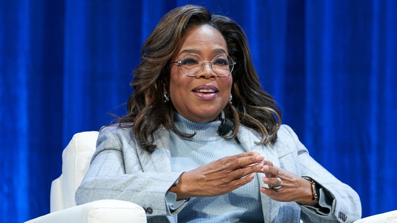 Oprah Winfrey sitting on a couch in a light blue suit and pointing her fingers