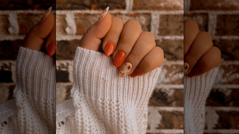 Closed hand showing a nail set with solid orange nails and two biege accent nails featuring fall leaves