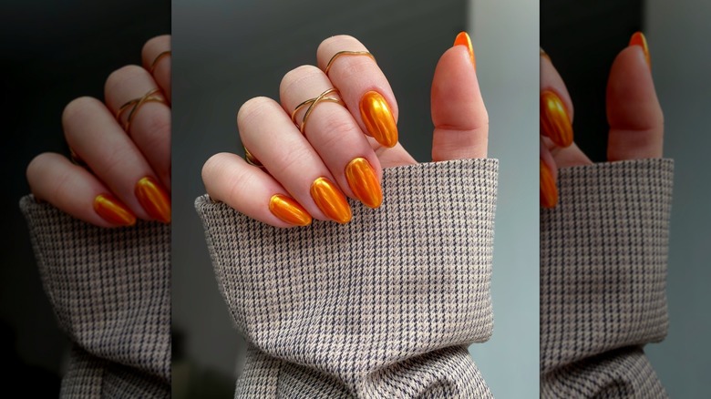 Mostly closed-hand showing orange nails with a chrome finish alongside a sleeve with a tweed pattern