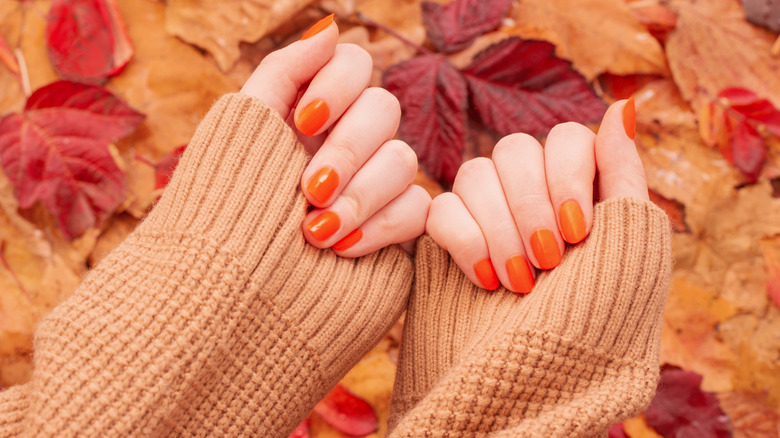Folded hands showing an orange nail polish alongside a beige sweater and fall leaves in the background