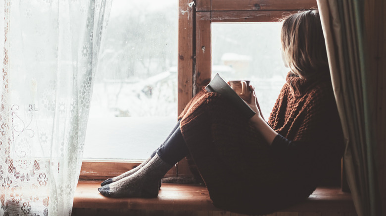 Young, cozy-looking person looking out on snowy landscape while sitting with book and mug on a windowsill