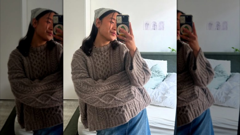 A woman taking a selfie in the mirror while wearing a brown cable-knit sweater and a bandana on her head